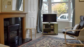 A living room with a fireplace, a television on a stand, a patterned rug, and a view of the outside through large windows. - 2 bedroom accommodation in Buxton (Buxton)