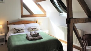 Une chambre avec un plafond en pente, un lit avec une literie verte, deux oreillers ornés de motifs d’oiseaux et une chaise dans un coin.