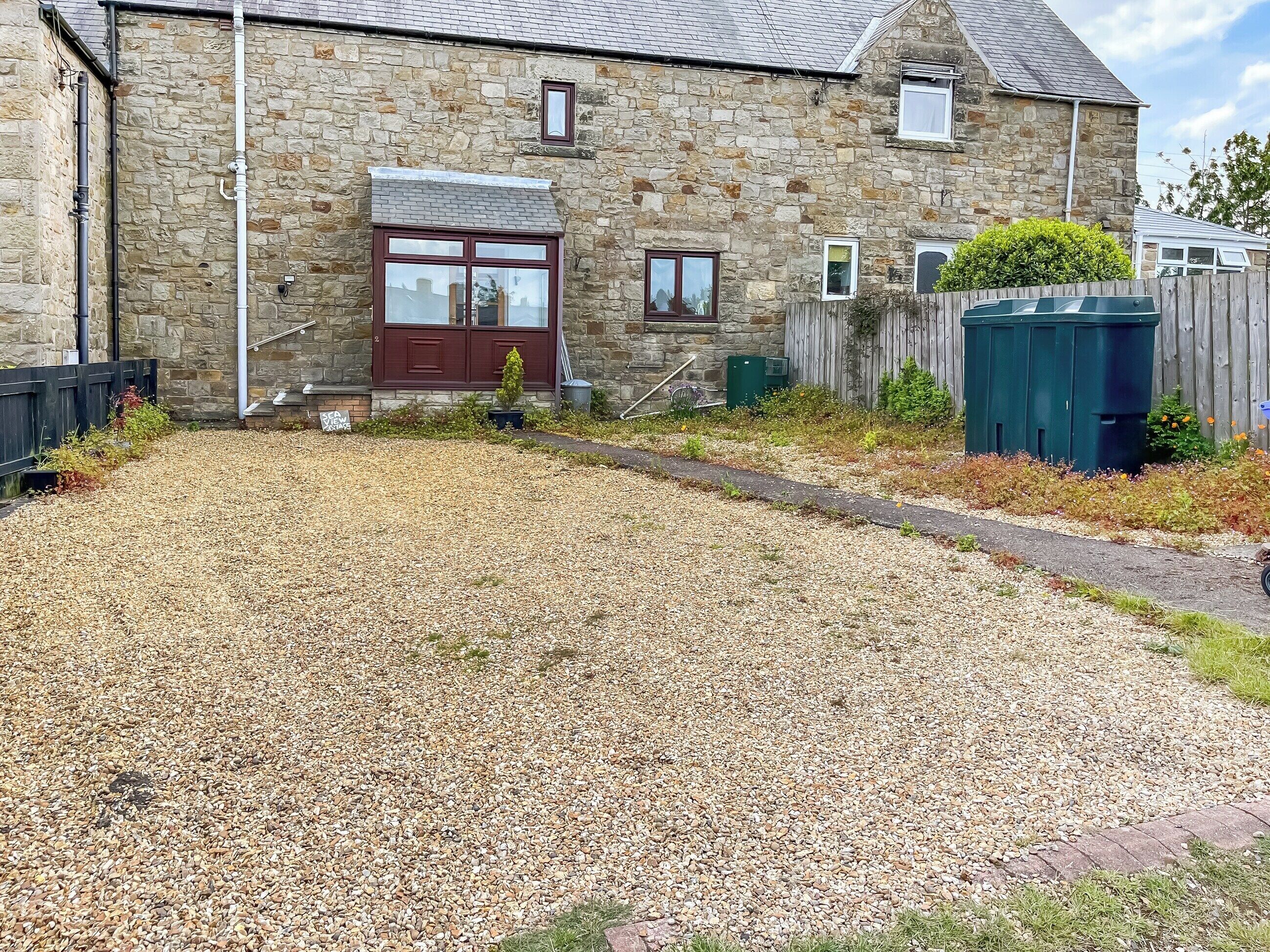 A stone building with a gravel driveway, a wooden fence, and a green bin.