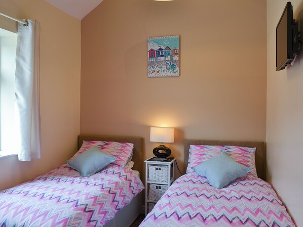 Une chambre avec deux lits, chacun recouvert d’une couvre-lit à motifs et agrémenté d’un oreiller. On y trouve un téléviseur fixé au mur et une lampe sur une table de chevet.