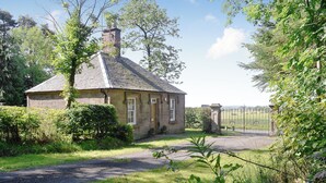 Een stenen huis met een pannendak, omgeven door weelderige begroeiing en een hekwerk.