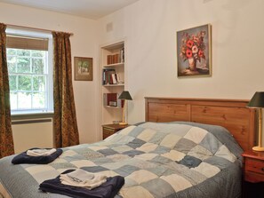 Ein Schlafzimmer mit einem hölzernen Bettgestell, einer gesteppten Bettdecke, einem Nachttisch mit Lampe, einem Bücherregal und einem Fenster mit Vorhängen.