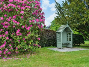 A small, light green wooden structure with a triangular roof, surrounded by a well-maintained lawn and a large bush with pink flowers.