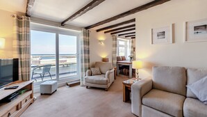 Een gezellige woonkamer met een televisie, bank en fauteuil, een eethoek met een tafel en stoelen, en een balkon met uitzicht op de zee.