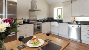 A modern kitchen with stainless steel appliances, a wooden dining table with a plate of food and wine glasses, and a window with a view of greenery. - 3 bedroom accommodation in Ventnor (Ventnor)