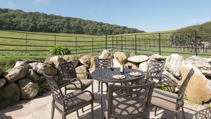 An outdoor patio with a table and chairs, surrounded by rocks and a view of a green field and hills. - 3 bedroom accommodation in Ventnor (Ventnor)
