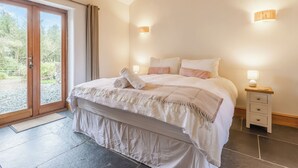 A bedroom with a large bed, two bedside tables, a sliding glass door to a balcony, and a stone-tiled floor. - The Cart House - HNNH (Hartland)