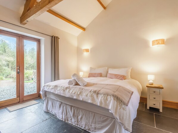 A bedroom with a large bed, two bedside tables, a sliding glass door to a balcony, and a stone-tiled floor. - The Cart House - HNNH (Hartland)