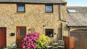 Una casa de piedra con puerta de madera, dos ventanas, un pequeño porche con banco y un jardín lleno de flores rosas.