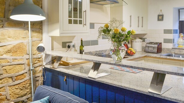 Una cocina con armarios blancos, pared de piedra, encimera azul y un jarrón con flores.