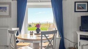 Una habitación con una mesa redonda de madera, sillas blancas, un jarrón con flores, un cuadro en la pared y vistas al exterior a través de una ventana con cortinas azules.