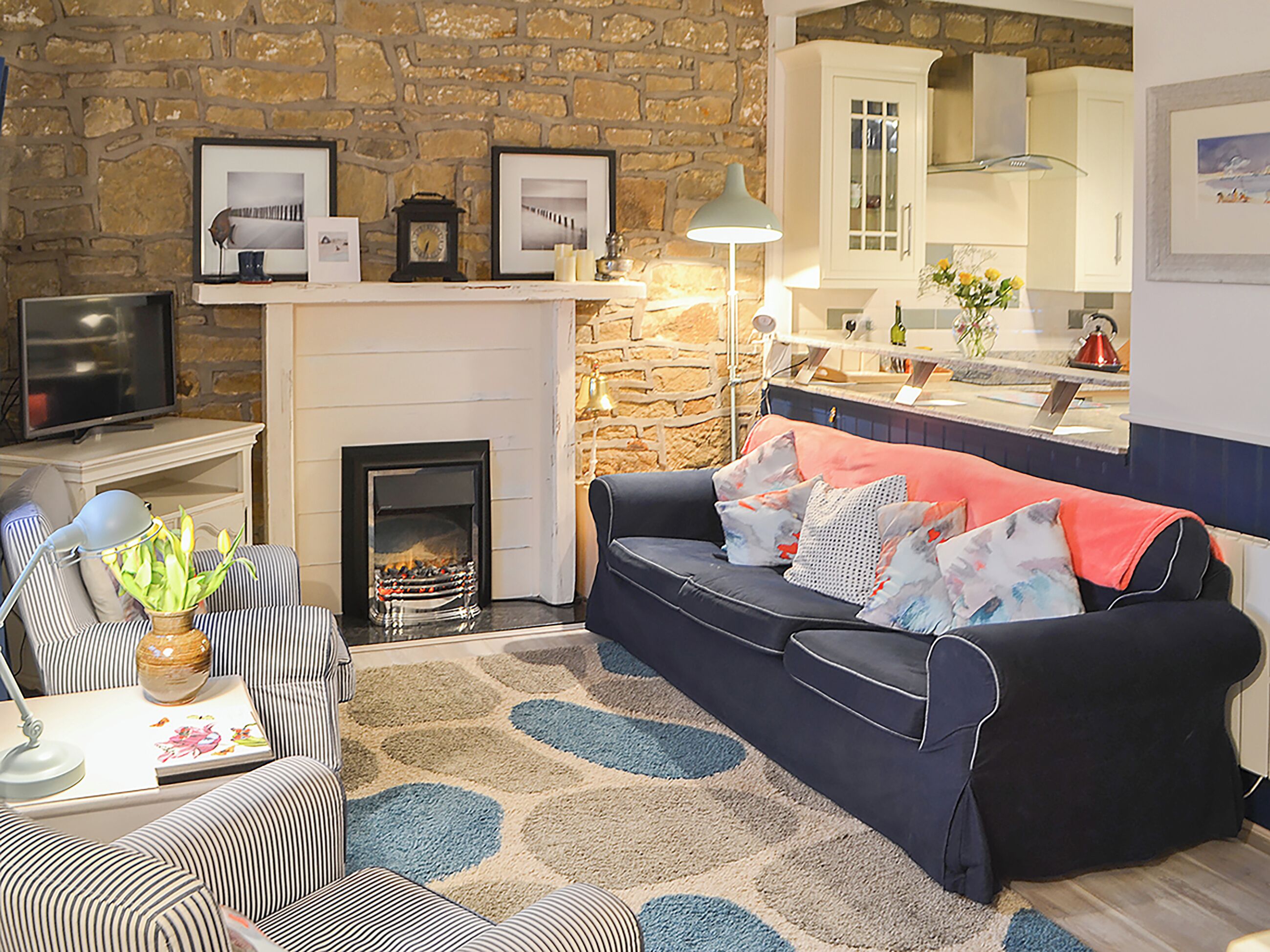 A cozy living room with a stone wall, a fireplace, a sofa with patterned cushions, a striped armchair, and a television.