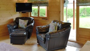 Ein gemütliches Blockhaus-Interieur mit zwei Ledersofas, einem Wand-Fernseher und großen Fenstern mit Blick ins Freie.