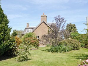 A stone house with a sloped roof and a chimney, surrounded by a well-maintained garden with various shrubs and flowering plants. - Islabank Farmhouse - SAAP (Meigle)