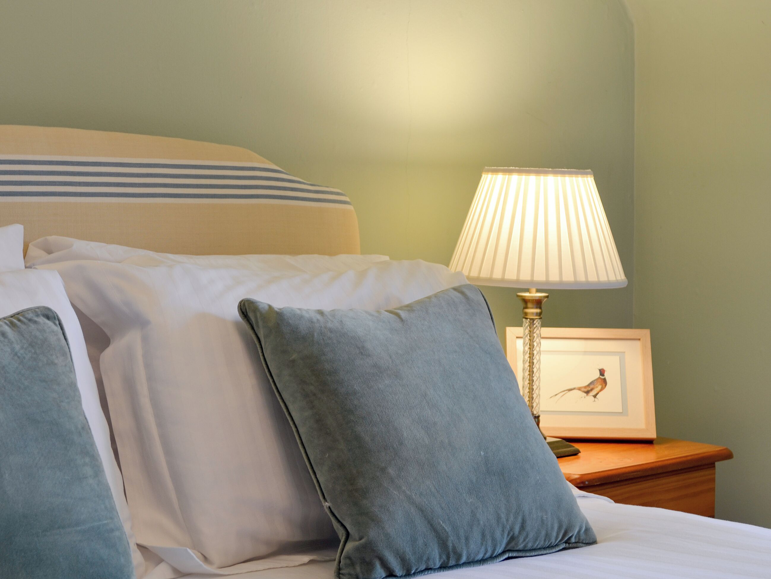 A neatly made bed with white and blue bedding, a blue pillow, and a bedside table with a lamp and a framed picture.