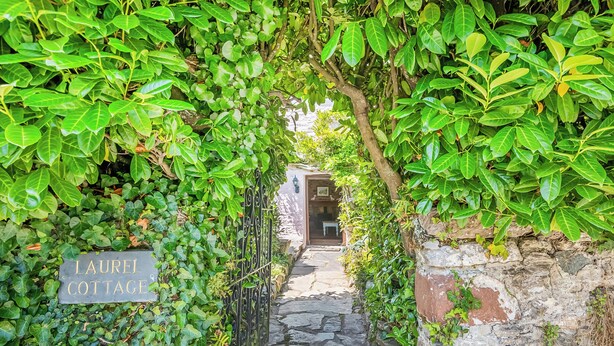 Un stretto sentiero in pietra conduce a un cottage con la scritta 'LAUREL COTTAGE', immerso nel verde e circondato da un rustico muro in pietra.