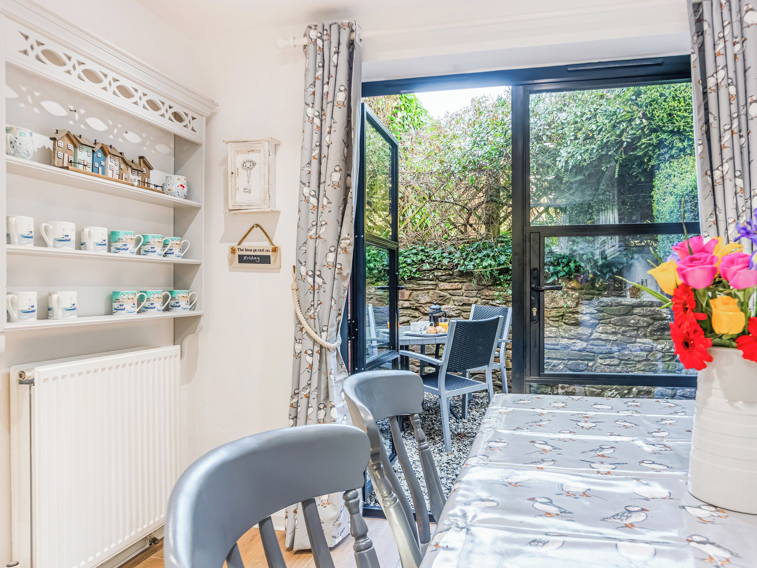 Un comedor con una mesa dispuesta para dos personas, un radiador, estantes con tazas y vistas a un jardín a través de una puerta de cristal.