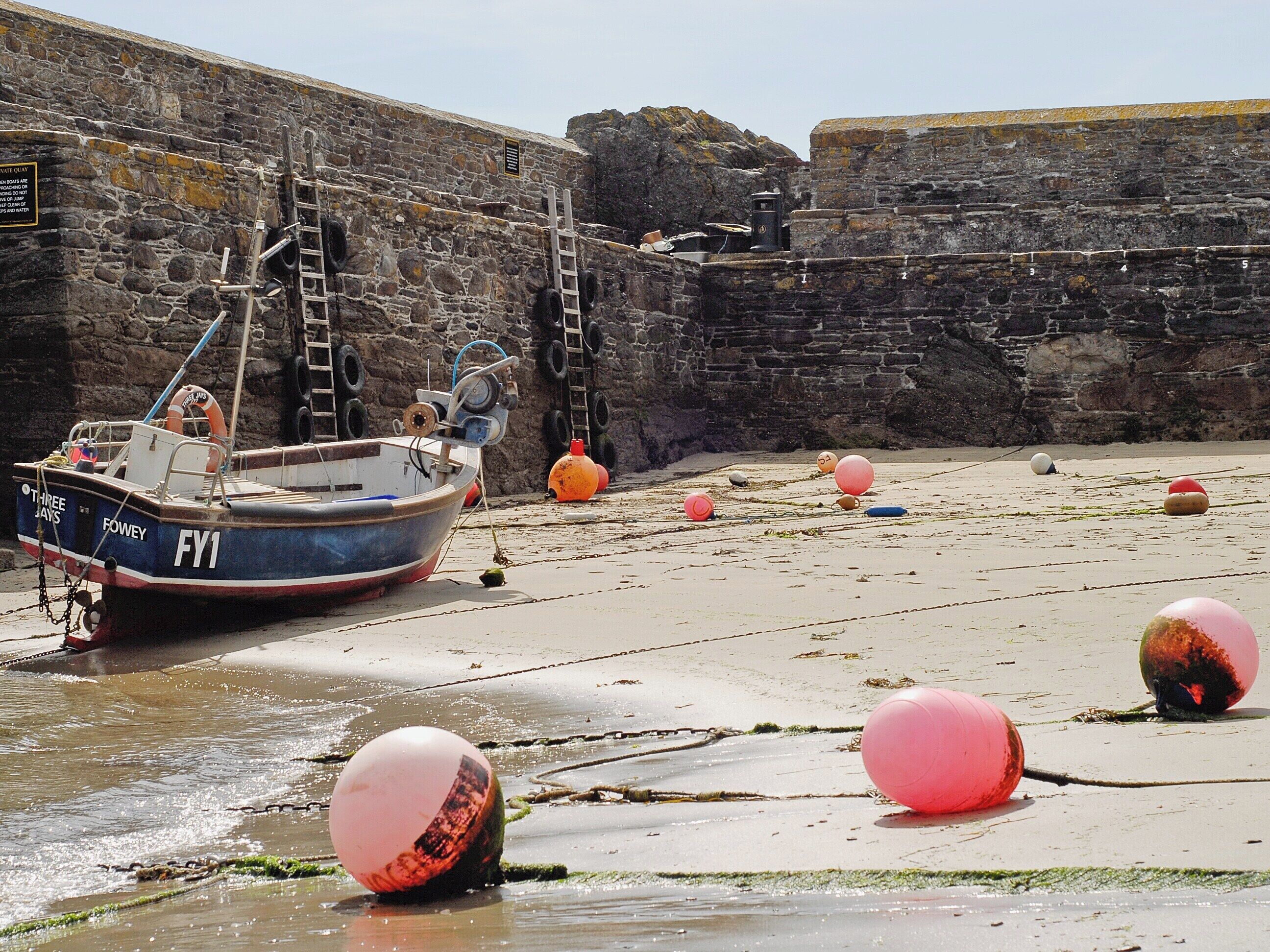 A stone wall with a boat named 'FY1' docked at a sandy beach with several orange buoys.