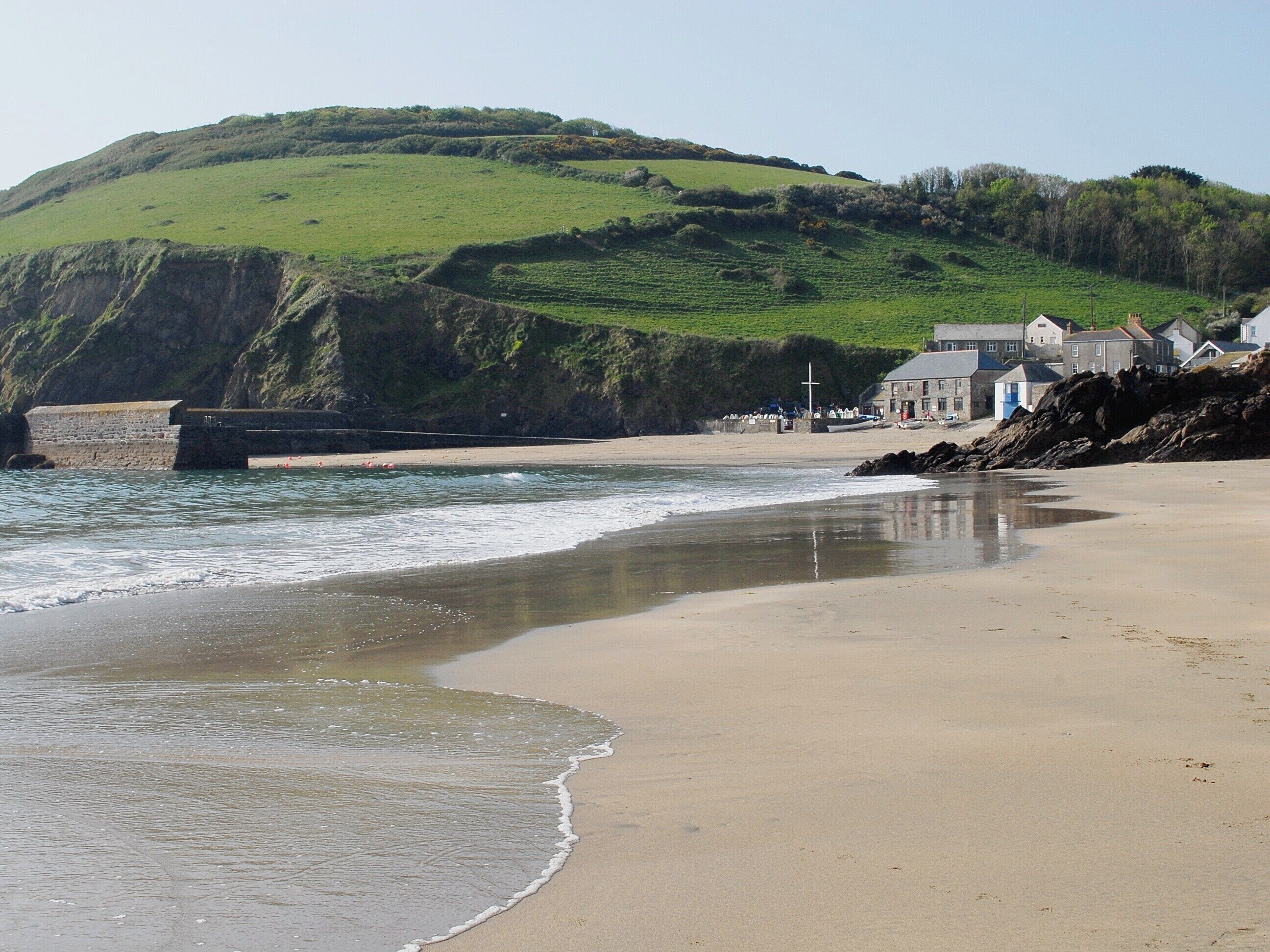 A coastal beach with a stone sea wall, houses, and a hill in the background.