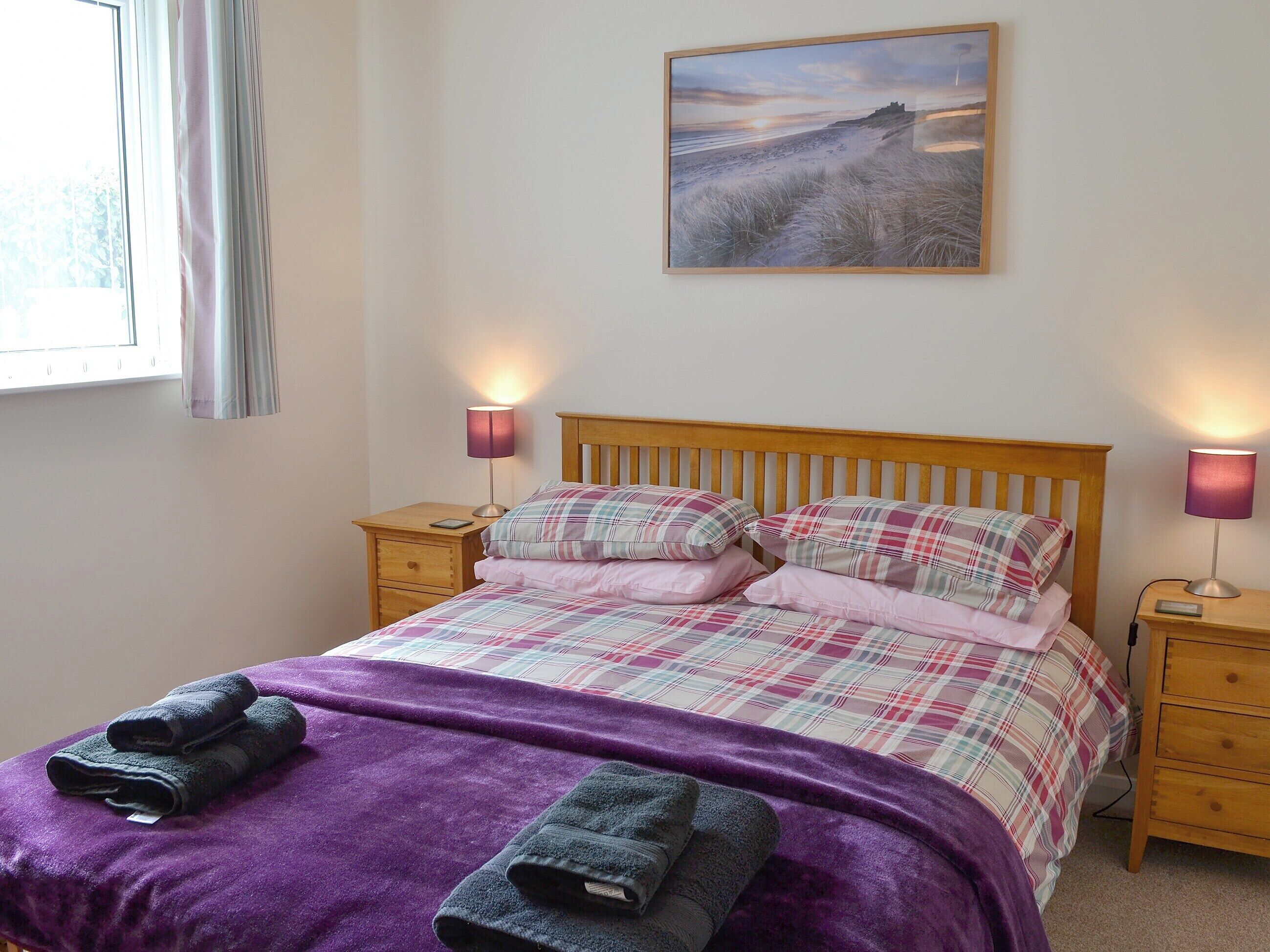 A bedroom with a bed, two bedside tables, a window with curtains, and a painting on the wall.