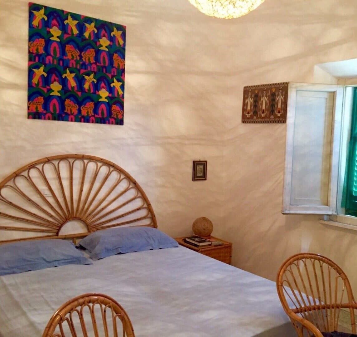 Un dormitorio con una cama, dos sillones de mimbre, una mesita de noche con un libro y una lámpara, un adorno en la pared y una ventana con cortinas.