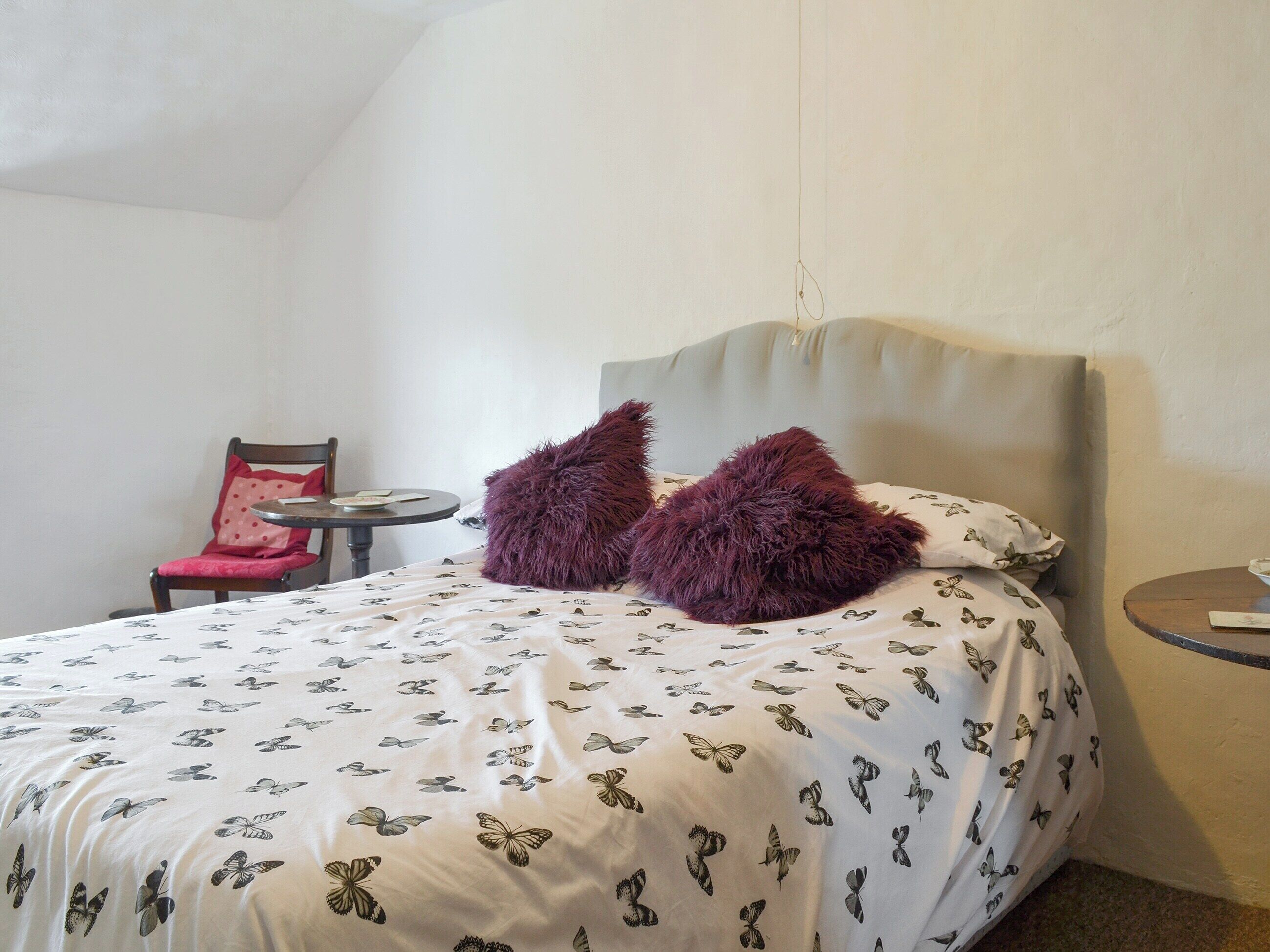 A bedroom with a bed featuring a butterfly pattern bedspread, a round side table, a chair with a red cushion, and a headboard.