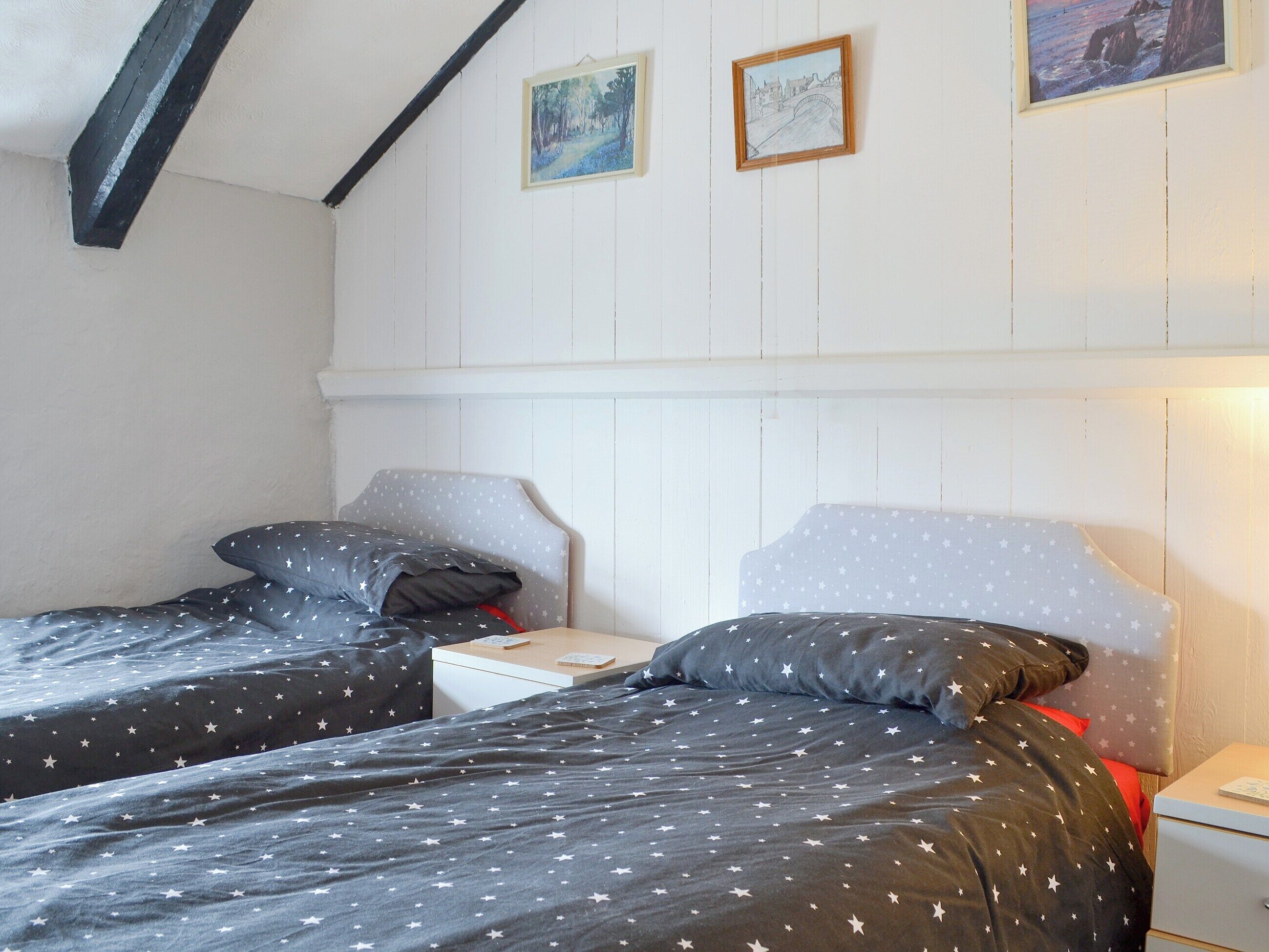 Ein Schlafzimmer mit zwei Einzelbetten, jeweils mit Kopfstütze und Kissen. An einem Nachttisch steht eine Lampe, auf dem anderen ein Buch. Die Wände sind weißes Wandverkleidung und die Decke ist schräg mit freiliegenden Balken.