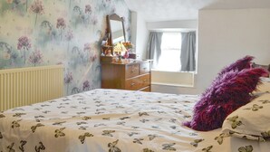 Een slaapkamer met bloemenbehang, een bed met een motief van vlinders, een dressoir, een spiegel en een raam met gordijnen.