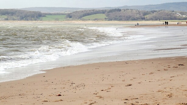 Une plage avec des vagues, une rive sablonneuse et des collines à l’horizon.