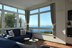 Una habitación con un sofá azul, dos mesitas redondas y vistas al mar a través de amplios ventanales.