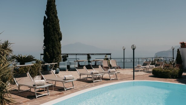 Zona junto a la piscina con sillones de descanso, un árbol alto y vistas al mar y a las montañas.