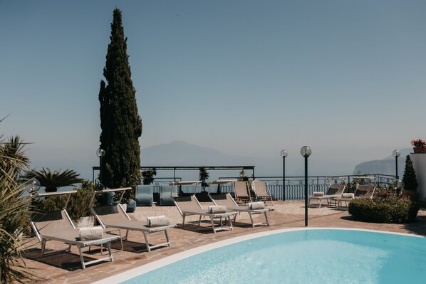 A poolside area with lounge chairs, a tall tree, and a view of the sea and mountains.