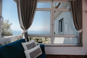 Ein Zimmer mit großem Fenster, einem blauen Sofa und Blick auf Meer und Berge.