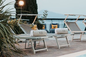 A poolside area with lounge chairs, some with towels, and a large tree in the background.
