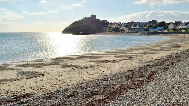 A beach with pebbles and seaweed, a castle on a hill, and houses in the background. - Single storey barn conversion just a mile from a sandy beach. (Criccieth)