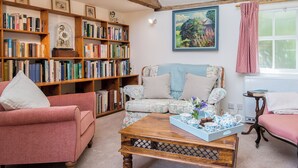 Ein gemütliches Wohnzimmer mit einem blumigen Sofa, einem Holztisch, einem Bücherregal voller Bücher und einem Bild an der Wand.
