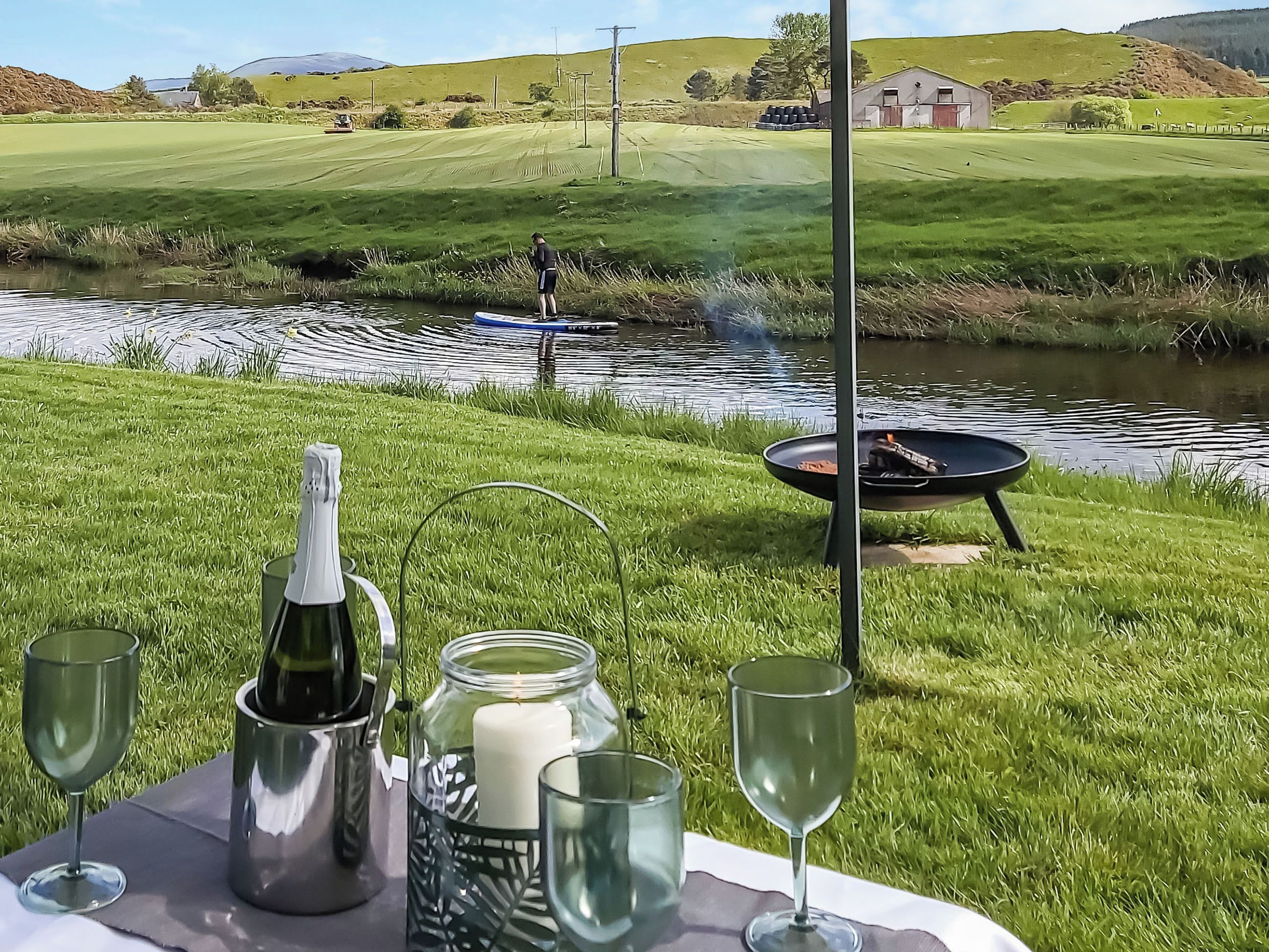 A picnic setup with a bottle, candle, and glasses on a table, overlooking a grassy field and a body of water.