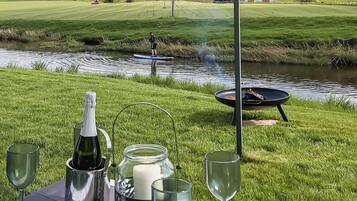 A picnic setup with a bottle, candle, and glasses on a table, overlooking a grassy field and a body of water.