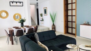 Uma sala de estar moderna com um sofá escuro, uma área de jantar com mesa branca e uma cozinha com armário de madeira.