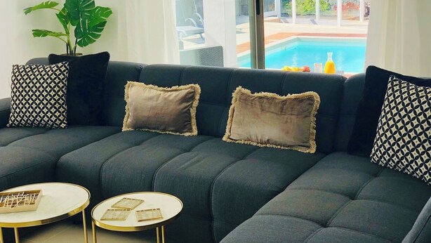 Uma sala de estar moderna com um sofá cinza escuro, almofadas estampadas e duas pequenas mesas redondas. Uma grande janela oferece vista para uma piscina e área externa.