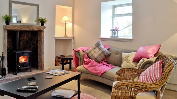 A cozy living room with a fireplace, a sofa, a wicker chair, and a table with remote controls and a book. - 3 bedroom accommodation in Gartocharn, near Drymen (Gartocharn, near Drymen)