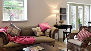 A living room with a sofa, a coffee table, a wicker chair, a lamp, and a window with a view of greenery. - 3 bedroom accommodation in Gartocharn, near Drymen (Gartocharn, near Drymen)