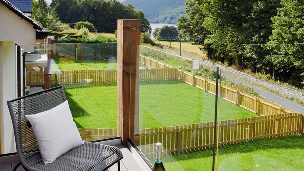 A balcony with a chair, a table, a bottle of wine, and a view of a green lawn and wooden fence. - 3 bedroom accommodation in Balnaskeag, near Kenmore (Balnaskeag, near Kenmore)
