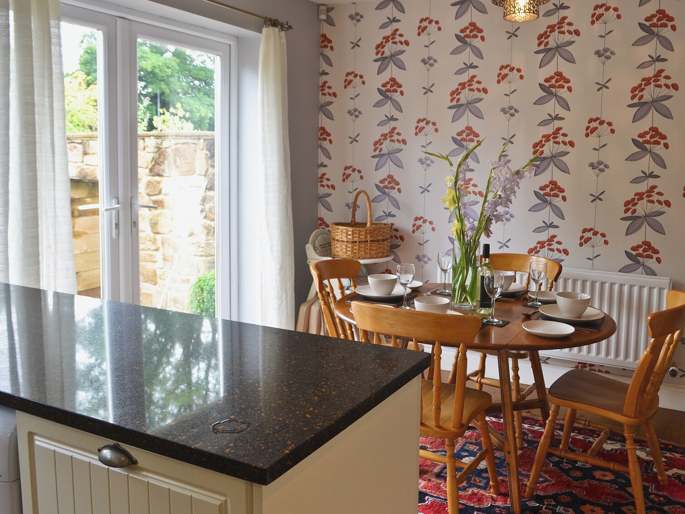 A dining area with a round table set for four, surrounded by wooden chairs. A black countertop extends from the wall, and a patterned wallpaper decorates the walls.