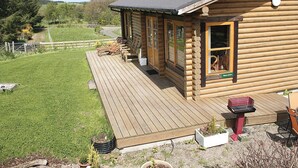Une cabane en rondins dotée d’une terrasse, de meubles de jardin et offrant une vue sur un espace verdoyant bordé d’arbres.
