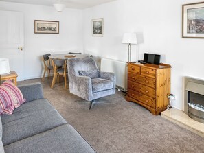 Un salon avec un canapé gris, un fauteuil à motifs, une commode en bois, une table avec des chaises et une cheminée.