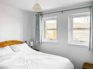 Une chambre à coucher avec un lit, deux fenêtres, un rideau et une table de chevet avec une lampe.