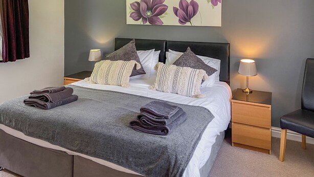 A neatly made bed with a grey blanket and folded towels, a headboard with multiple pillows, a bedside lamp, and a wall-mounted artwork of purple flowers. - Looking for a romantic escape in Herefordshire’s rolling countryside. (Tarrington, near Ledbury)