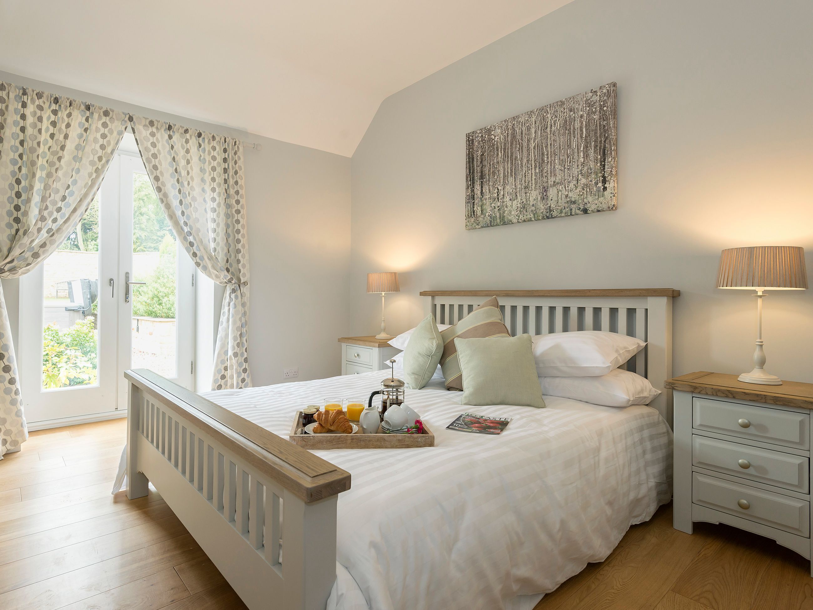 A bedroom with a bed, bedside tables, a window with curtains, a painting on the wall, and a tray with breakfast items.