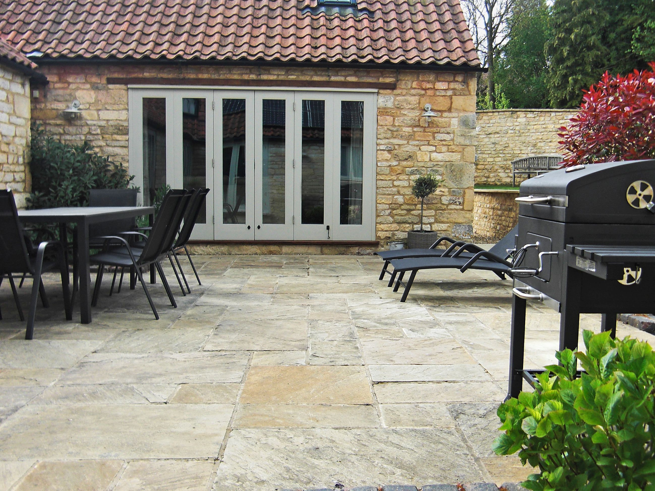 A patio with stone flooring, a table with chairs, a grill, and a stone wall with a window.